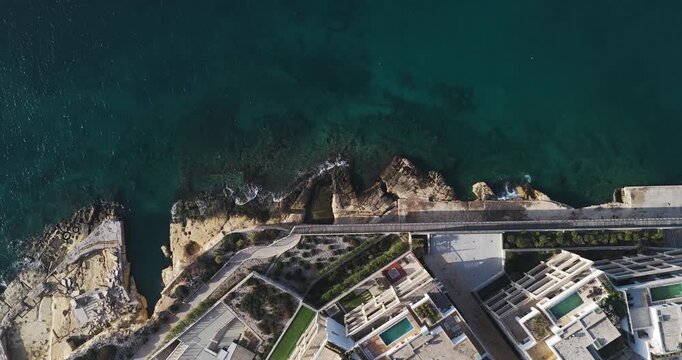 Aerial view of the rocky coastline meeting modern buildings with pools, showcasing a contrast of natural textures and geometric shapes, Sliema, Malta.