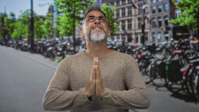Man with hands pressed together in prayer gesture, wearing glasses, bare hands visible, looking up on an amsterdam street beside rows of parked bicycles; hope faith.