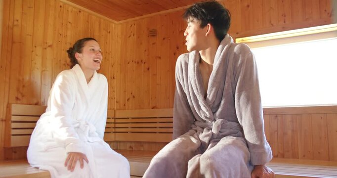 Diverse couple sitting on bench in sauna starting chat, turning, laughing and relaxing in robes