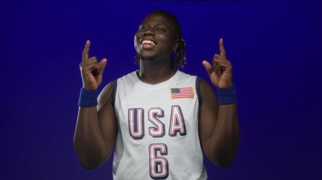 Man pointing index fingers upward while smiling, wearing white usa basketball jersey with flag patch and blue wristbands in blue studio; pride.