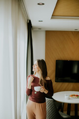 Obraz premium Woman stands by window holding cup in modern room with light decor and furniture