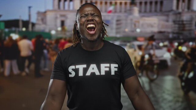 Young african american man wearing staff shirt with open mouth shouting and hands raised, dreadlocks visible in crowded city street; passion urban energy.