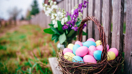 Obraz premium A wicker basket overflows with colorful Easter eggs against a weathered wooden fence. Lilies frame the scene.