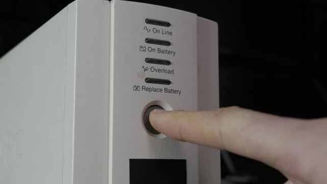 A finger presses the power button on a UPS battery unit, activating the yellow indicator light. Concept of data protection, emergency power supply, and electrical maintenance.