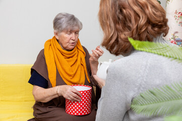 Elderly woman opening a red polka dot gift box while another woman offers a white teapot. Generational care and gift giving concept