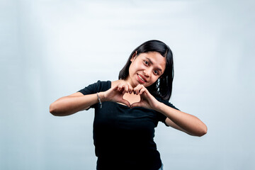 Fototapeta premium Cheerful Indonesian woman in a black t-shirt forming a heart symbol with her fingers and smiling, isolated on white background.