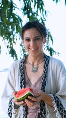 A beautiful brunette in a bohemian style relaxes outdoors in the summer, holding a large slice of watermelon against a green forest backdrop.