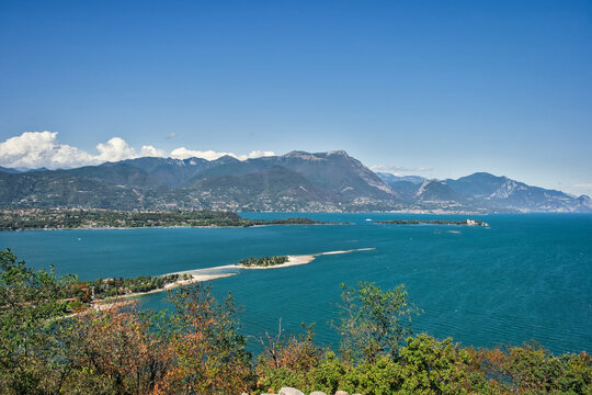 High-angle vista of vibrant turquoise waters and distant mountains under a clear blue sky, viewed from the cliffs of Rocca di Manerba, Italy.