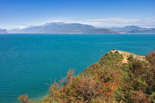 High-angle vista of vibrant turquoise waters and distant mountains under a clear blue sky, viewed from the cliffs of Rocca di Manerba, Italy.