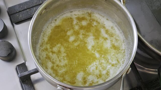 Yellow melted butter bubbles and foams inside a stainless steel saucepan on a gas stove burner. Concept of homemade cooking, clarifying butter, and classic dairy preparation.