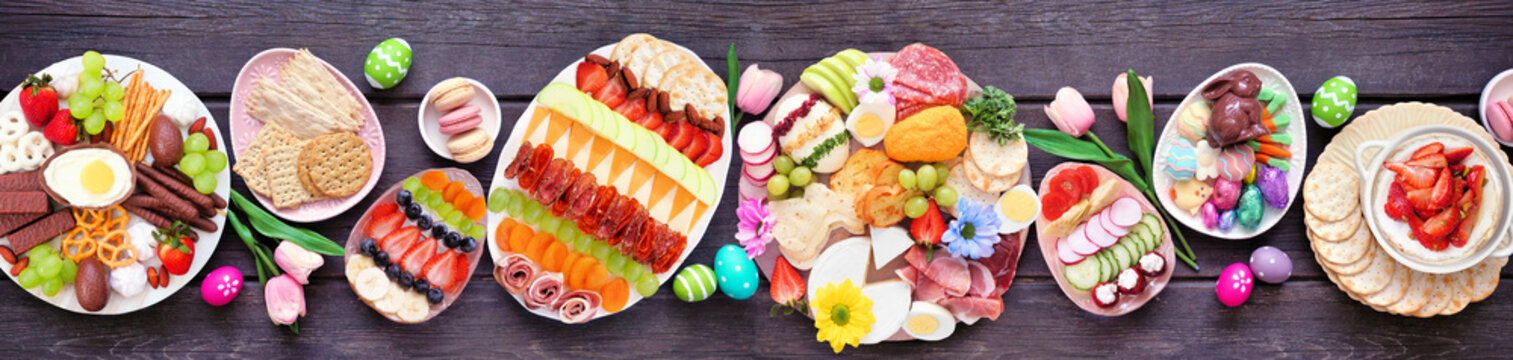 Spring or Easter grazing board theme table scene against a dark wood banner background. Overhead view. Assortment of sweets and cheese, meat and fruit appetizers.