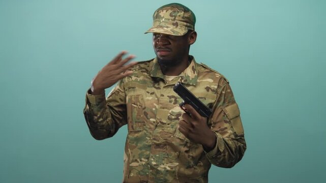 Soldier in camouflage uniform holding pistol pinches nose with thumb and forefinger while standing against a studio teal backdrop under flat light; stress duty.
