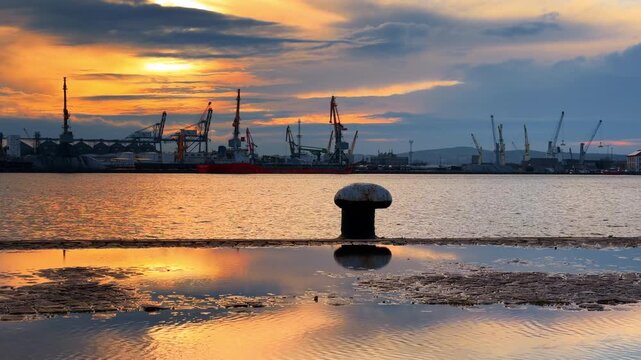 Large mooring bollard on the dock shore near a huge puddle. Skyline of container cranes booms at backdrop. View on the port at sunset.