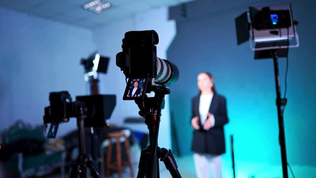 Approaching cameras on tripods and soffits working in photo studio. Woman stands at blurred backdrop talking for blog content.