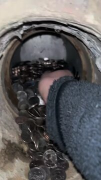 Hand reaching through a circular metal opening into a large pile of mixed coins during collection sorting or machine maintenance showing practical currency handling and manual financial work