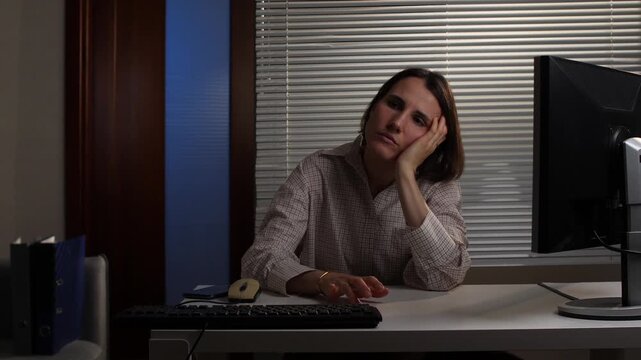 Woman sitting at a minimalistic office desk in a dimly lit room, resting her head on her hand, demonstrating exhaustion, stress, and overwork during late hours