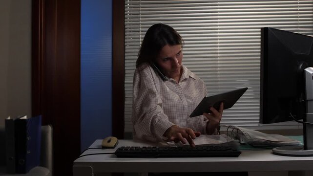 Businesswoman staying late in the office working on multiple tasks, talking on a smartphone, and consulting a digital tablet while sitting at her desk with a computer monitor
