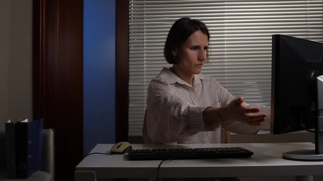 Woman focusing intently on a computer monitor, reaching out with an agitated gesture, indicating potential stress or a technical issue during her late night work session at the office