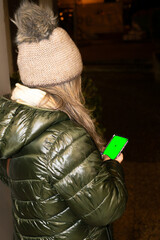 Woman wearing a green anorak and hat on the street holding a smartphone with a green screen for editing.