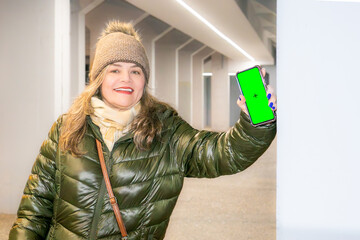 Woman wearing a green anorak and hat on the street holding a smartphone with a green screen for editing.