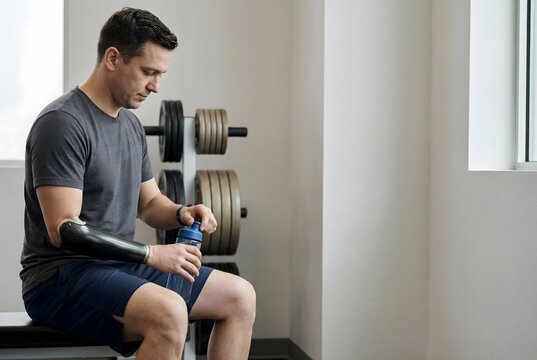 Caucasian man with prosthetic arm resting in gym, holding bottle. Para-athlete at training session in sport club, concept of disability sport.