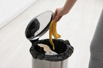 Woman throwing banana peel into trash bin in kitchen, closeup