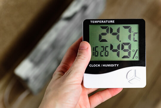 Hand checking room temperature and humidity on a digital thermometer near a heating radiator.