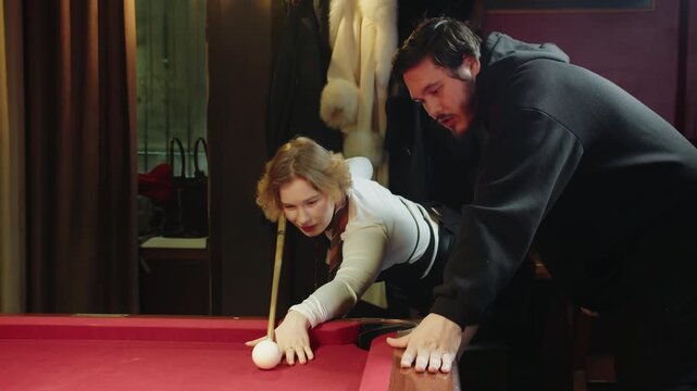 Young smiling female with cue enhances her billiards techniques under instructor supervision from adult caucasian solid man. Woman is taught how to play pool in cozy environment with warm lighting
