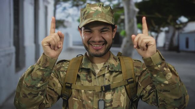 Hispanic soldier in camouflage uniform with visible dogtags points both index fingers upward to sky on street; pride duty hope.