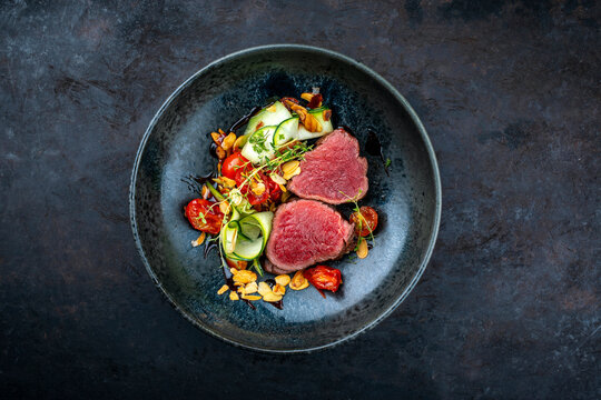Fine dinining dry aged beef filet steak rare tataki style cut tagliata with sliced zucchini, tomato and almonds served as top view in a design bowl