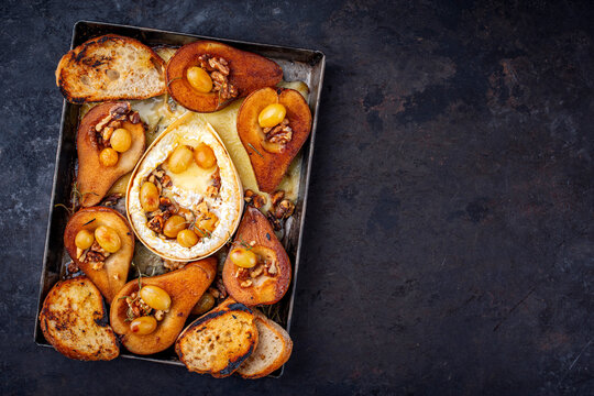 Oven baked camembert cheese with caramelized glazed pears, walnuts and grapes and baguette served on rustic metal plate