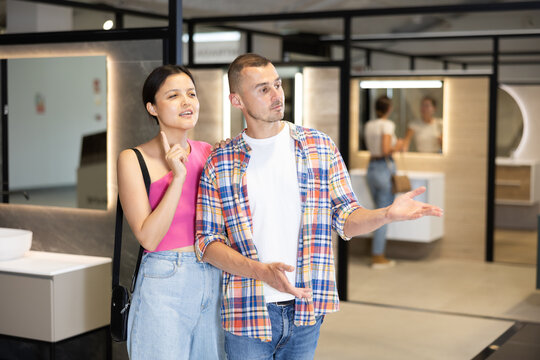 Interested young couple, man and woman of different ethnicities, amicably discussing choice of bathroom fixtures and furnishing during visit to specialized plumbing store..