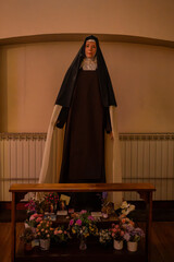Religious Statue of a Nun with Prayer Offerings and Flowers, Catholic Saint Figure in Traditional Habit with Devotional Altar, Interior Church View of a Saintly Statue Behind a Prayer Bench