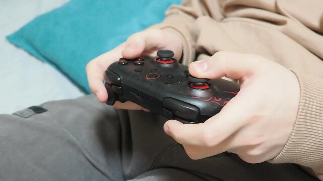 Concentrated boy in big gaming headphones playing video game with wireless controller console joystick, sitting on bed. Concept of gaming, children and technology, screen time, and leisure at holidays