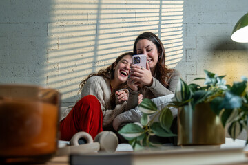 Two cheerful friends relaxing at home, smiling and laughing while watching something entertaining on mobile phone