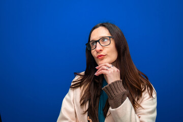 Woman wearing glasses with hand on chin, looking away, thinking and making a decision against a blue background