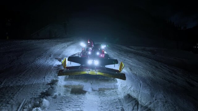 Drone close up of snow groomer with headlights working on ski slope at night. Grooming machine prepares fresh snow for winter resort activities.