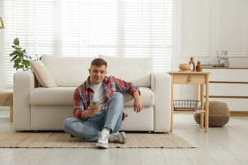 Young man with smartphone relaxing near sofa at home