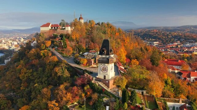 Aerial view of iconic Uhrturm clock tower on Schlossberg hill, beautiful autumn cityscape of Graz with red roofs and colorful trees. Graz at sunrise, view of Styria capital during fall season, Austria