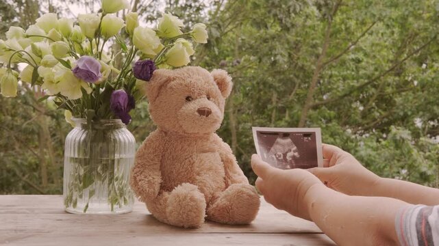 Ultrasound announcement card, sonogram image in hand, flowers in glass vase, Teddy bear on wooden table, pregnancy reveal, motherhood, baby coming soon, prenatal care, family joy, waiting for baby