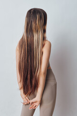 Obraz premium Woman with long straight hair standing with hands behind back wearing casual beige top and leggings in front of plain light background for beauty and style concept.