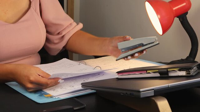 Close-up of a woman stapling papers at her desk. The image conveys office work, organization, productivity, and professional tasks in a modern workspace or home office environment.