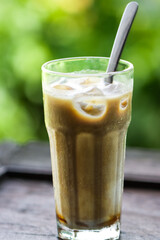 Refreshing Iced Latte Coffee with Milk Layers and Ice Cubes in Tall Glass on Dark Wooden Table, Summer Beverage with Natural Green Bokeh Background for Gastronomy and Outdoor Cafe Lifestyle Concept
