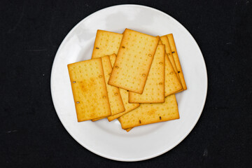 Fototapeta premium Crispy Wheat Crackers with Seeds on White Plate Top View, Savory Golden Brown Biscuits for Bakery and Culinary Branding, Healthy Crunchy Snack on Minimalist Black Background Flat Lay