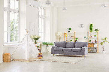 Home for a modern family. A bright minimalist living room interior with a gray sofa, white children's teepee tent, open shelving and green plants, offering a cozy family space and soft natural light © Studio Romantic