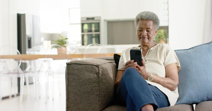 Senior African American woman sitting on sofa checking messages and scrolling smartphone at home