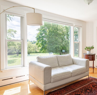 Bright modern living room with large picture windows and white sofa, sunlit interior overlooking green backyard landscape
