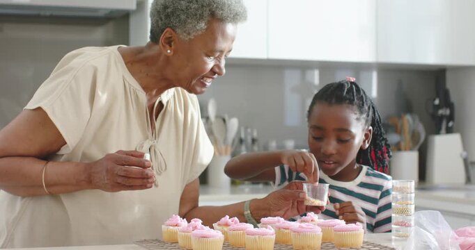 Senior woman offering sprinkles guiding youth granddaughter decorating cupcakes in kitchen