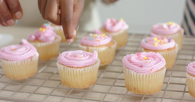 Hand entering frame is pinching small sprinkles and dropping onto pink cupcakes on rack for serving