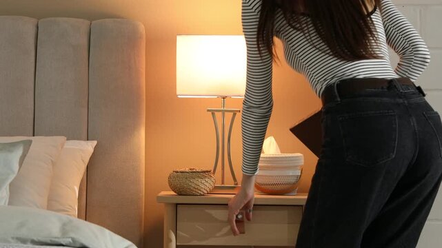 Woman putting book into bedside table drawer after lamp turning on at home